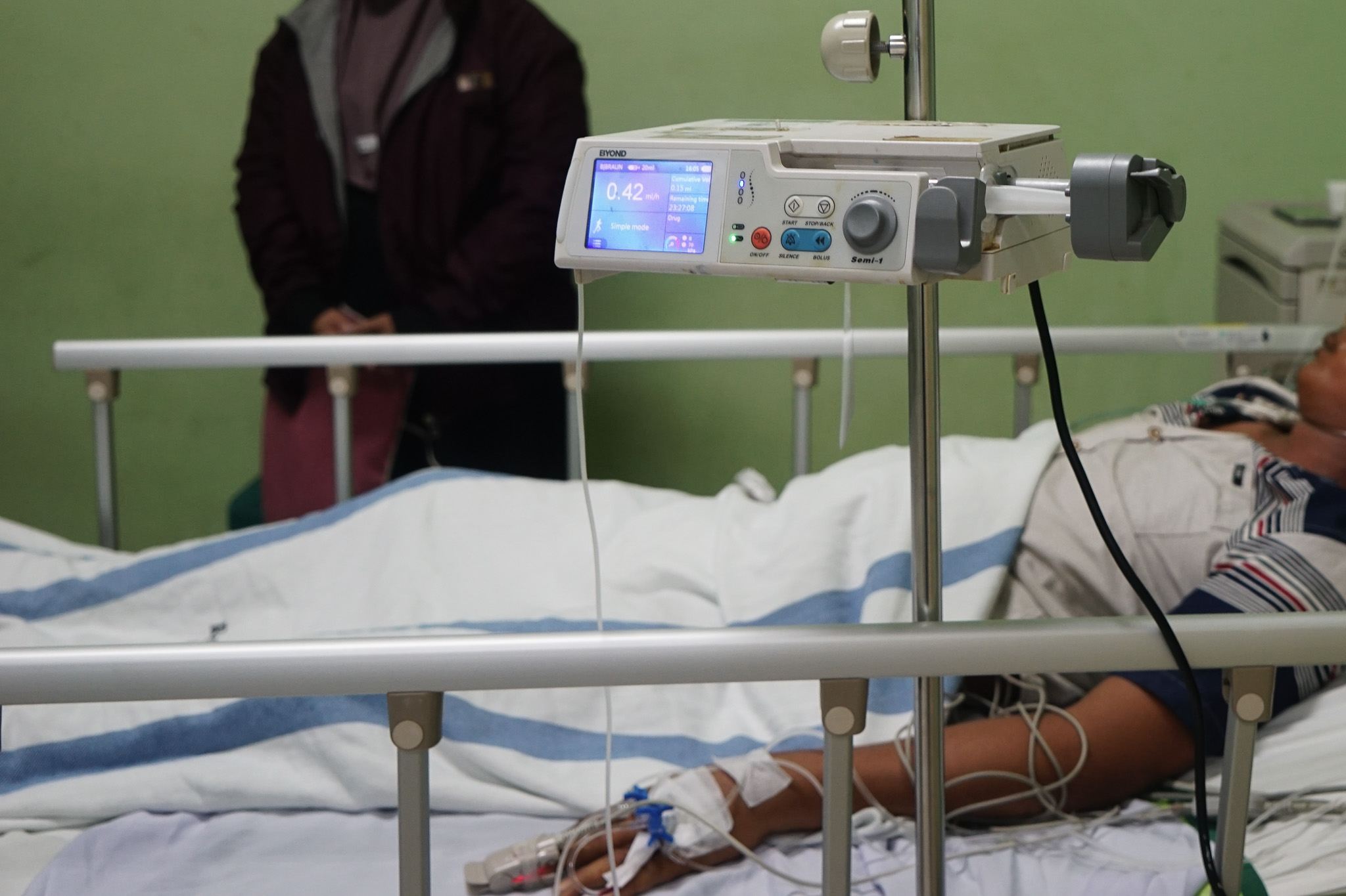 A patient lying in a hospital bed with medical equipment and a relative standing by their side.