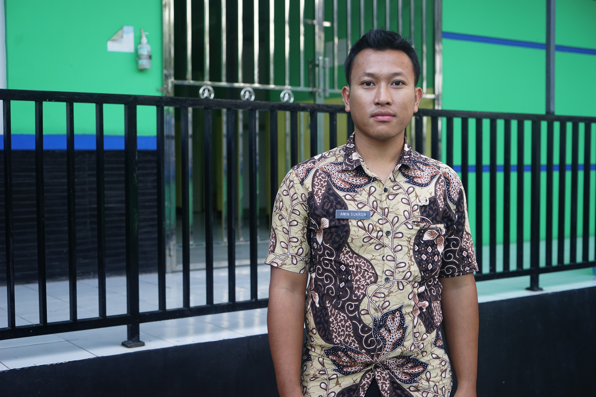 A person wearing a batik shirt with a name tag reading 'AMIN SURKON' stands in front of a green gate and wall.