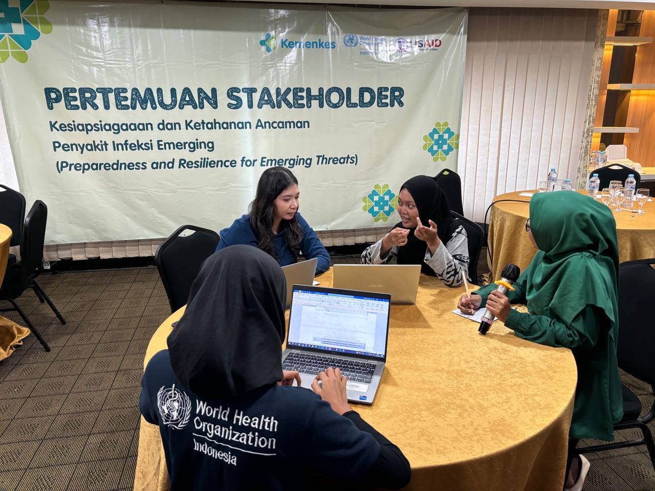 Four participants are sitting around a round table, discussing with a laptop and a microphone, taking notes.