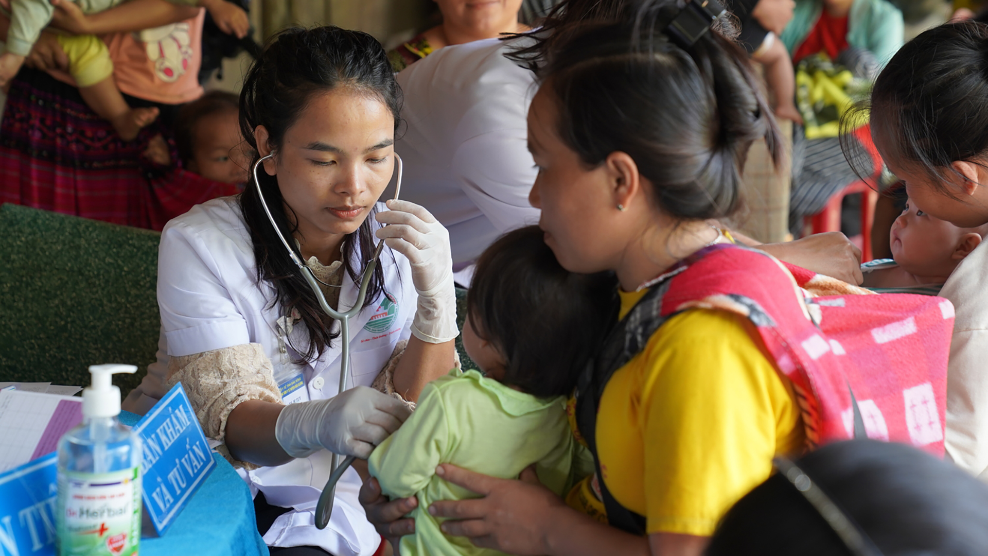 A female medical doctor is using a stethoscope to examine a child sitting in her mother's lap, in a busy environment with other mothers' and babies.